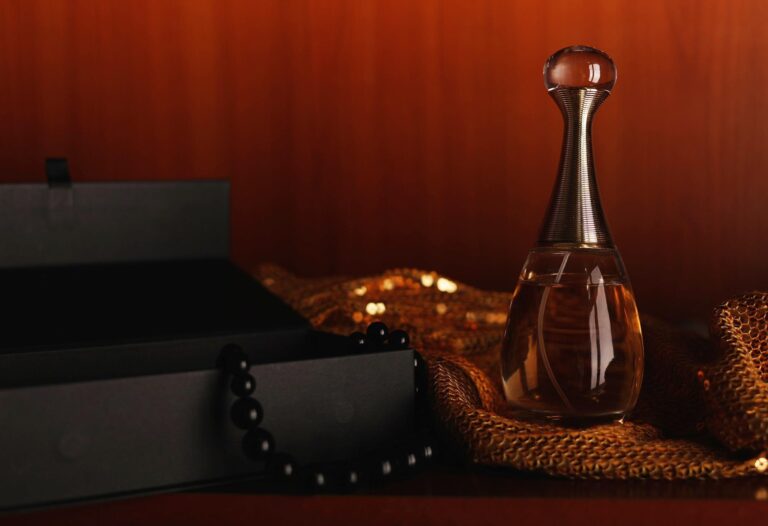 Fragrâncias orientais: o guia completo Frasco de perfume âmbar com tampa dourada sobre tecido de paetês dourados, ao lado de caixa preta e pérolas, com fundo de madeira quente