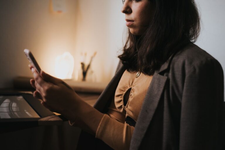 Mais de 40% das solicitações chegam fora do horário comercial e ampliam o tempo de resposta ao cliente Mulher de blazer verifica uma mensagem no celular em um ambiente com pouca luz, com o notebook aberto sobre a mesa ao lado