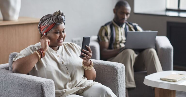 Como Escolher o Melhor Formato de Vídeo do Tiktok Para Assistir em Diferentes Dispositivos Mulher sorridente usando celular e homem com laptop em ambiente aconchegante